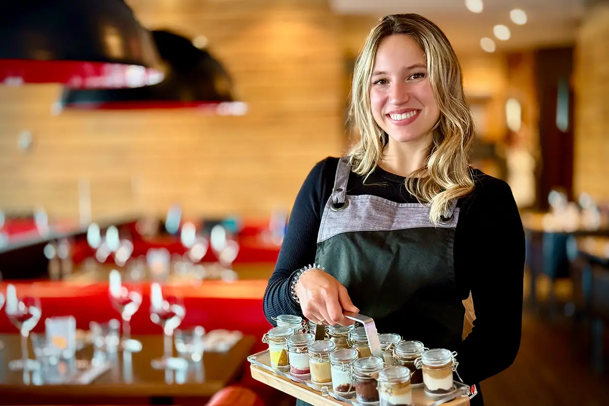 Employée de PACINI Alma présentant une sélection de desserts dans la salle à manger, illustrant le service attentionné et l’ambiance conviviale du restaurant.